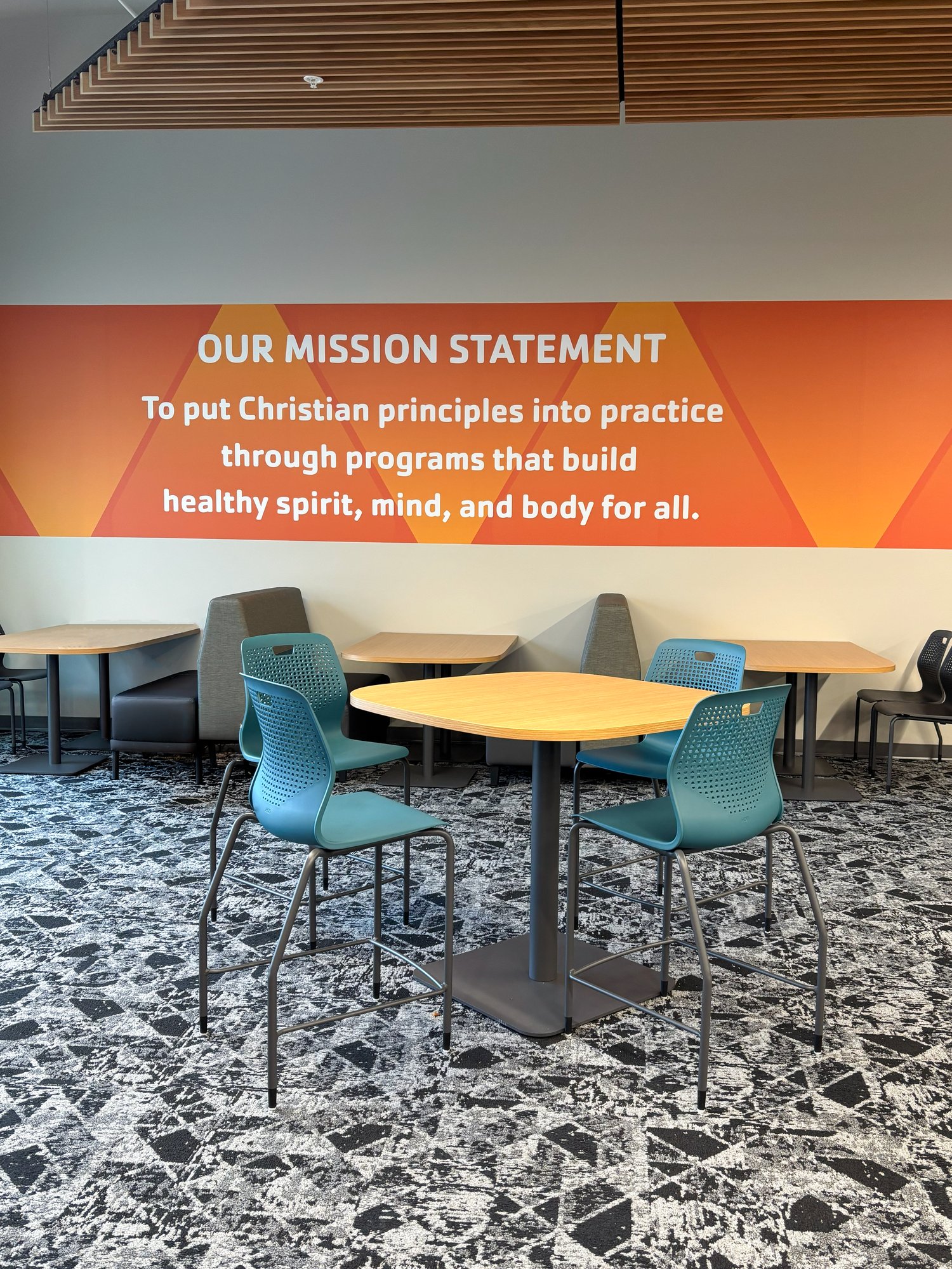 A table with four teal chairs sits in front of an orange mural with YMCA's mission statement on it A table with four teal chairs sits in front of an orange mural with YMCA's mission statement on it
