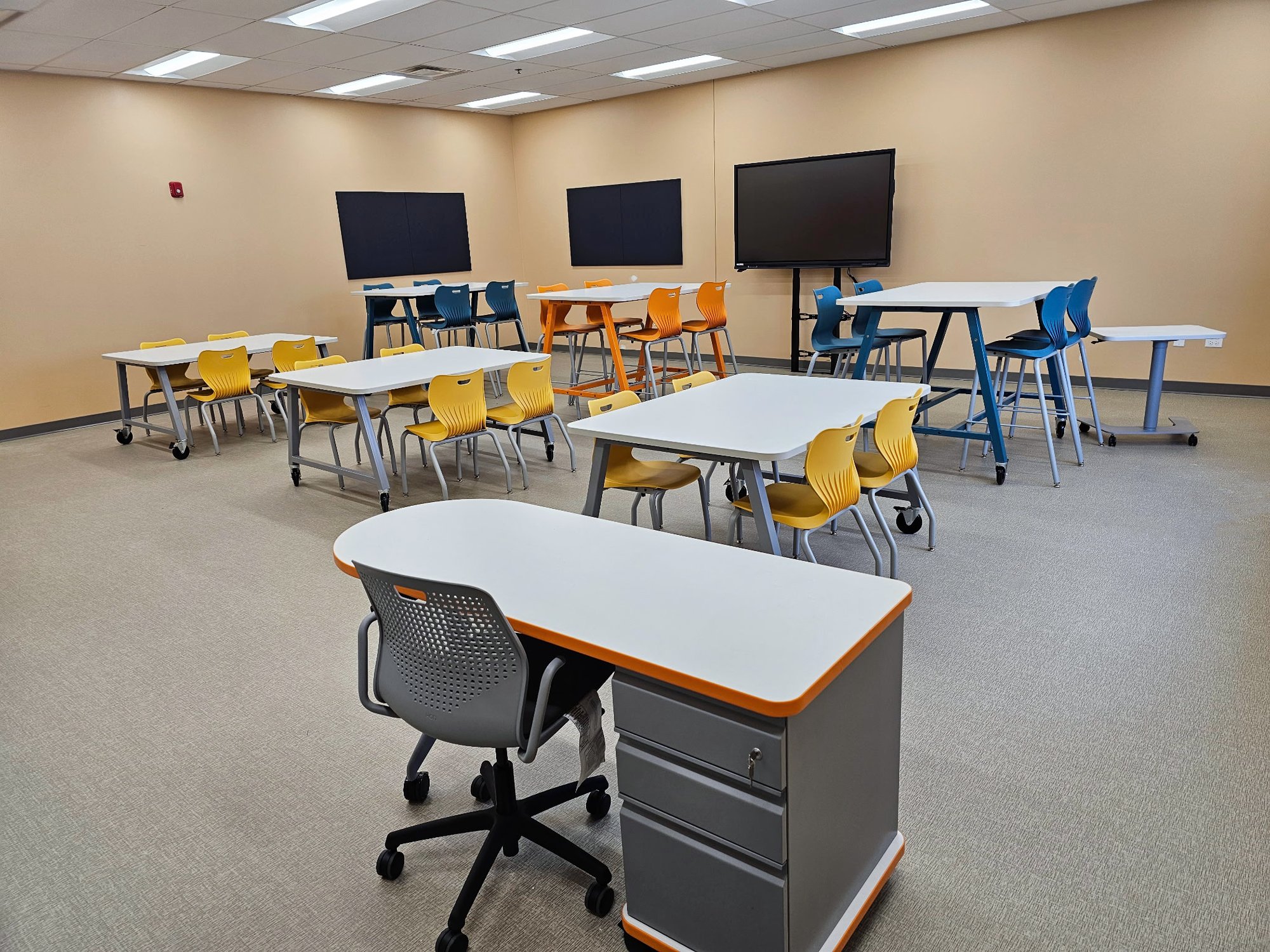 Classroom with various desks with yellow, orange, and blue chairs at each desk. Three TVs are mounted to the wall. Classroom with various desks with yellow, orange, and blue chairs at each desk. Three TVs are mounted to the wall.
