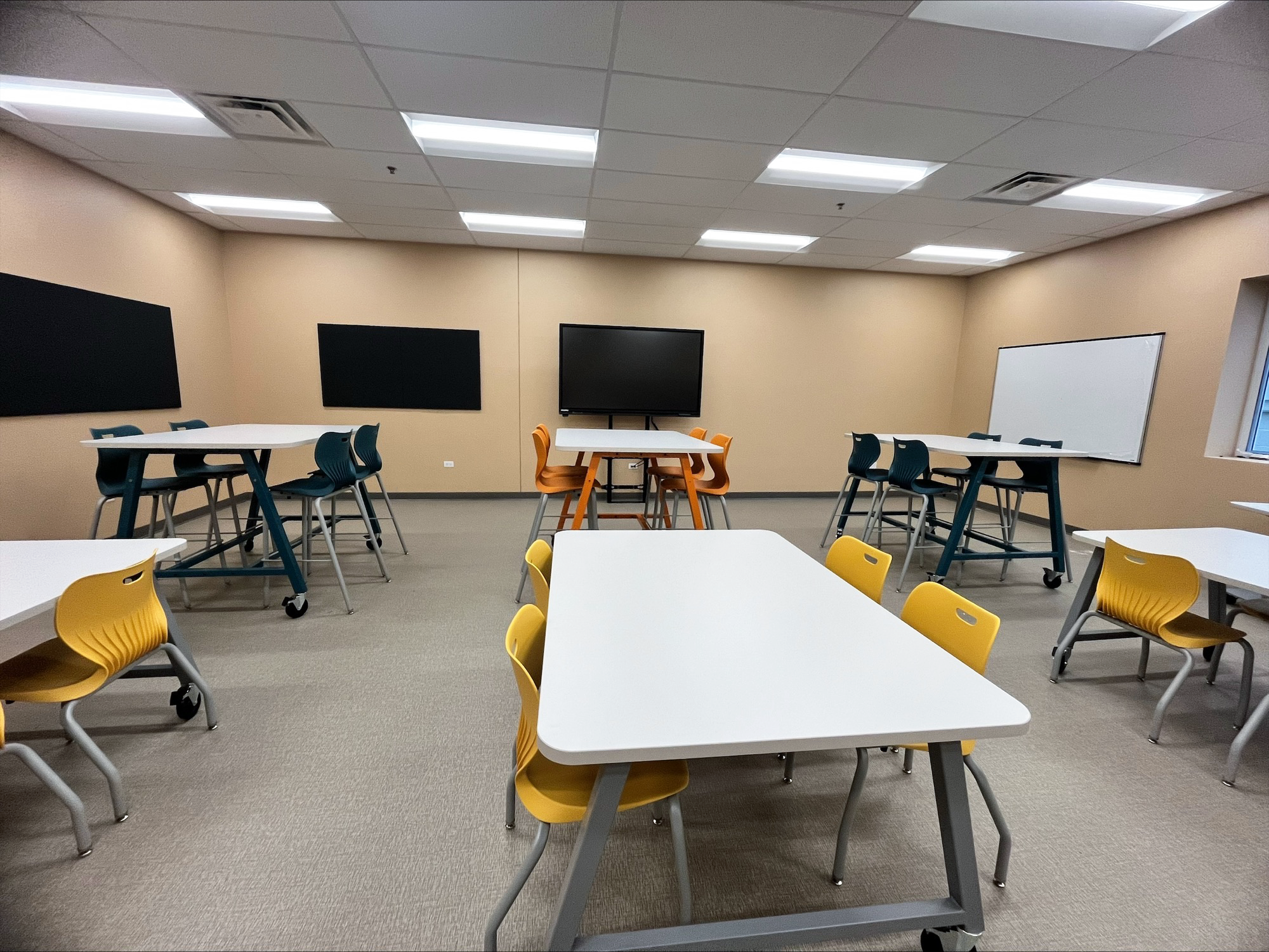 Six worktables are arranged in a classroom with blue, yellow, and orange chairs at them. Three TVs are mounted to the wall. Six worktables are arranged in a classroom with blue, yellow, and orange chairs at them. Three TVs are mounted to the wall.