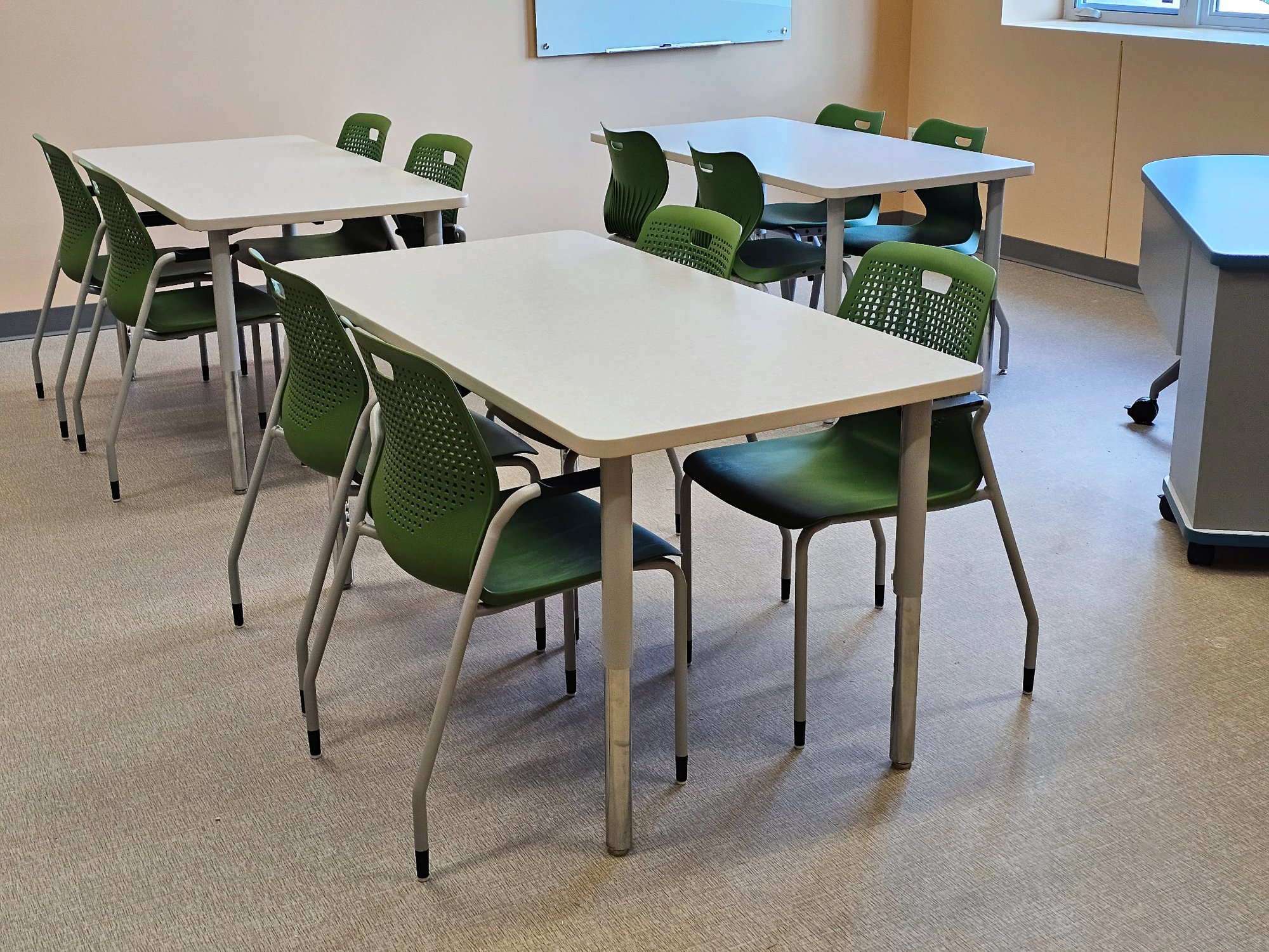 Three gray desks with green chairs sit in a classroom. Three gray desks with green chairs sit in a classroom.