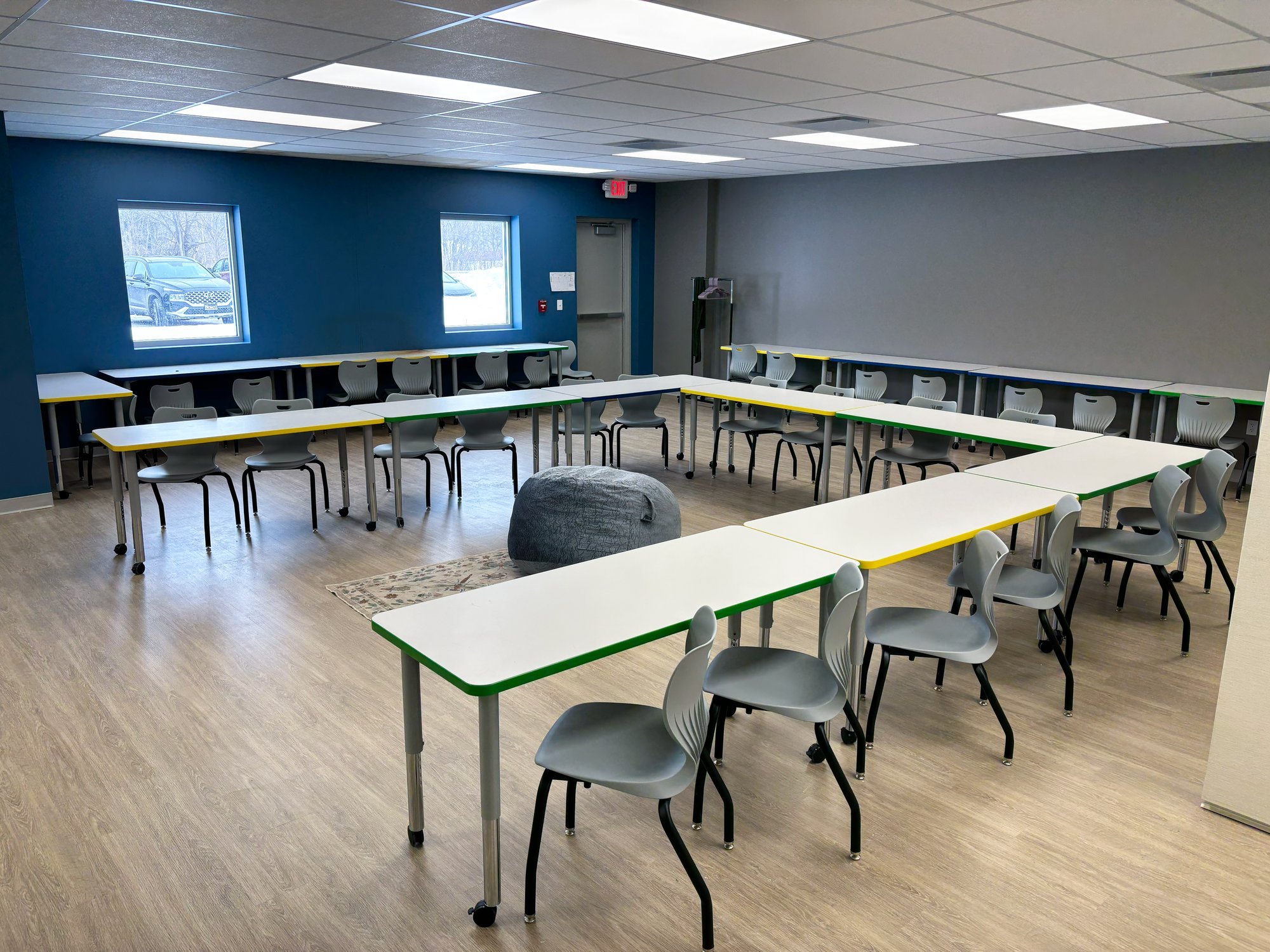 Tables with colorful edge banding are arranged in a "U" shape with chairs surrounding it. There's a large rug and bean bag chair in the center of the "U"