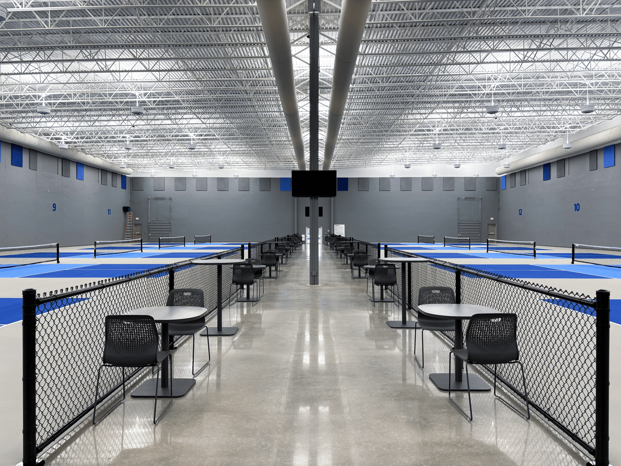 Pickleball courts line either side of a walkway lined with tables, chairs, and chain-link fencing Pickleball courts line either side of a walkway lined with tables, chairs, and chain-link fencing