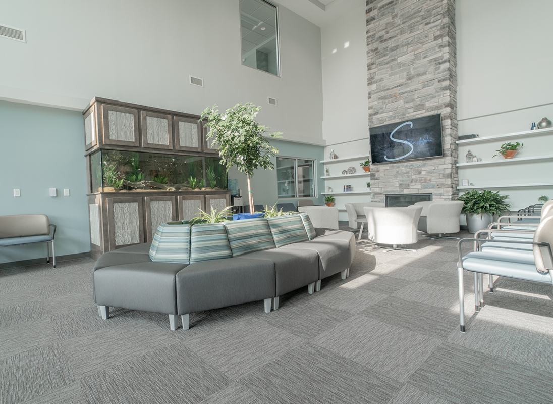 Couches sit in front of a large fish tank on the wall, with chairs and plush seating in front of a brick decorative fireplace