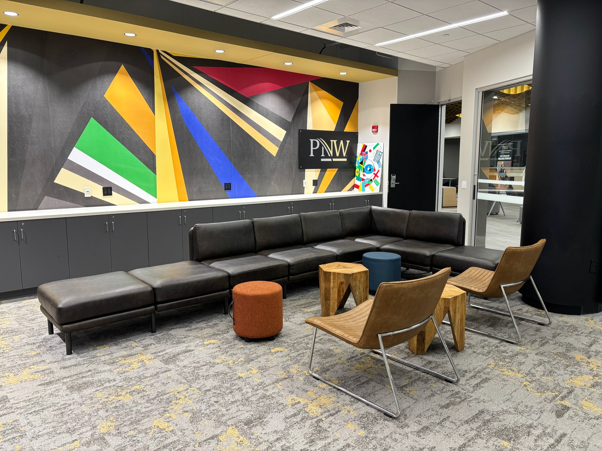 Lounge room with a multi-colored wall, a black, L-shaped couch, and tan chairs Lounge room with a multi-colored wall, a black, L-shaped couch, and tan chairs