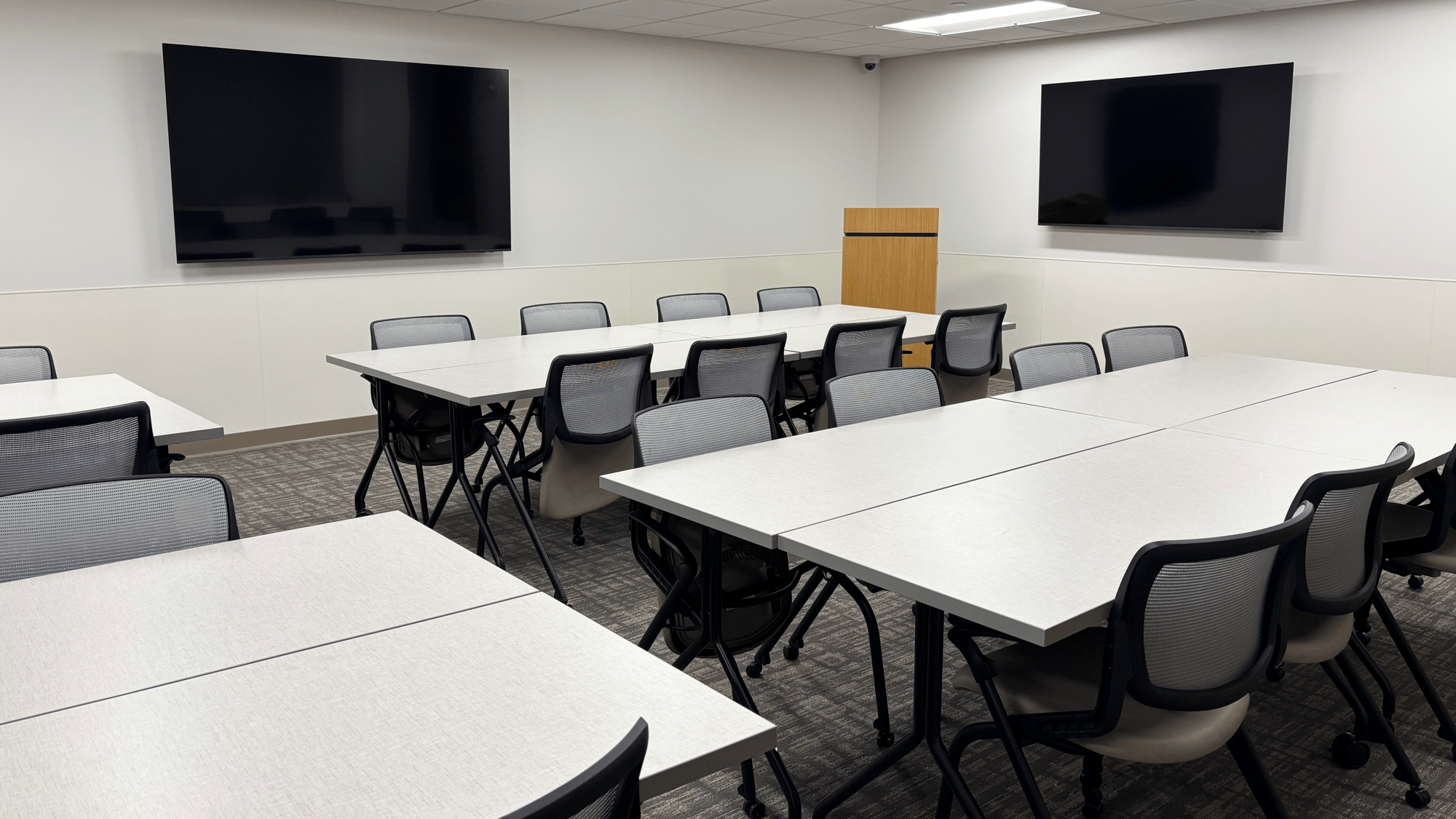 Training room with many tables, rolling chairs, and two TVs hung on the wall Training room with many tables, rolling chairs, and two TVs hung on the wall