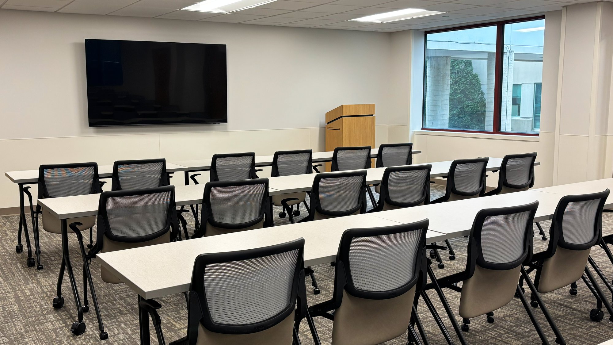 Rows of rolling chairs and tables with a wall-mounted TV and a podium Rows of rolling chairs and tables with a wall-mounted TV and a podium