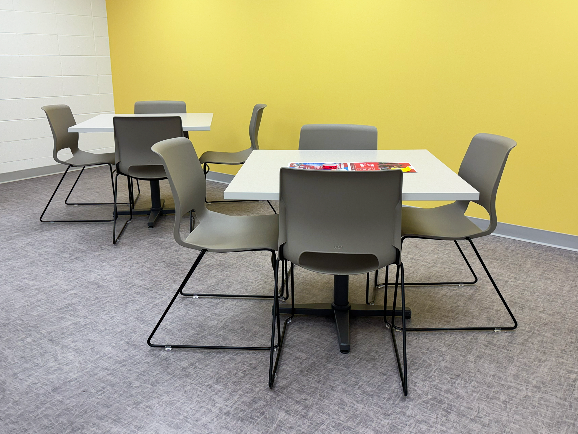 Two white tables are surrounded by grey chairs in front of a yellow wall