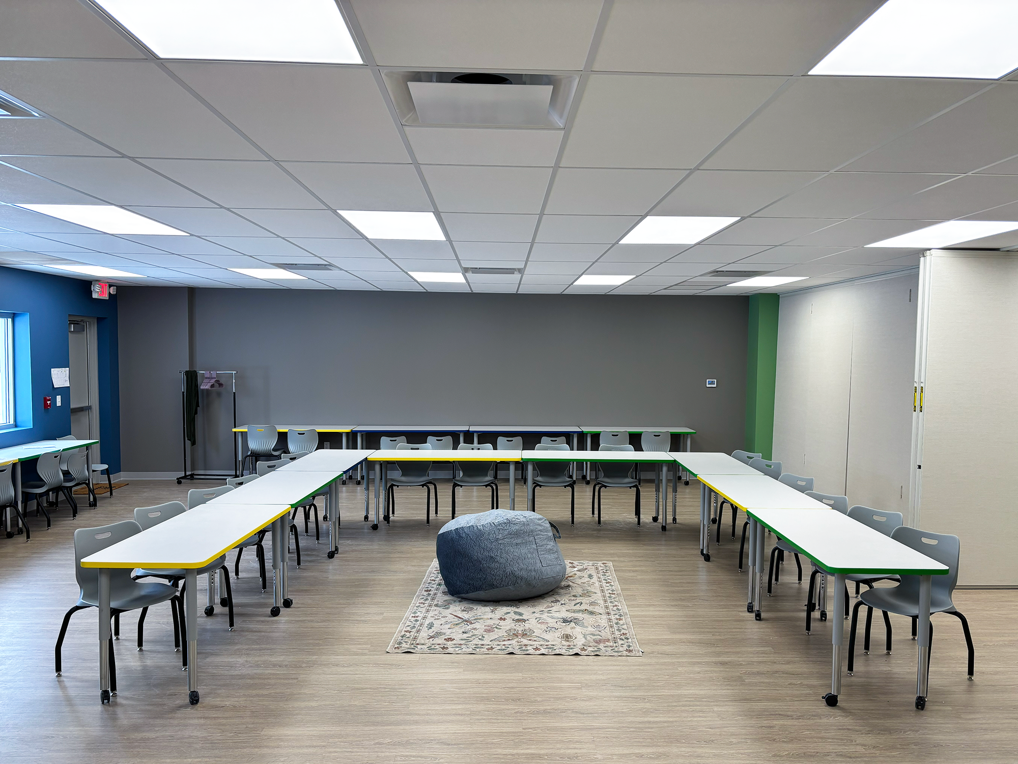 Tables with colorful edge banding are arranged in a "U" shape with chairs around it. There is a large rug and a bean bag chair in the center of the "U"