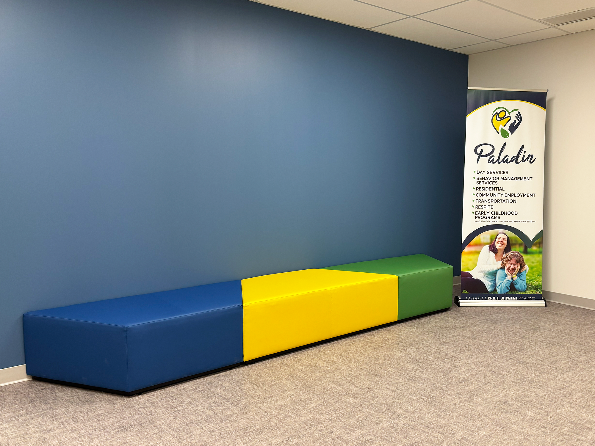 Blue, yellow, and green Hon Tangram sofas are pressed against a blue wall, with a Paladin banner sign to the right