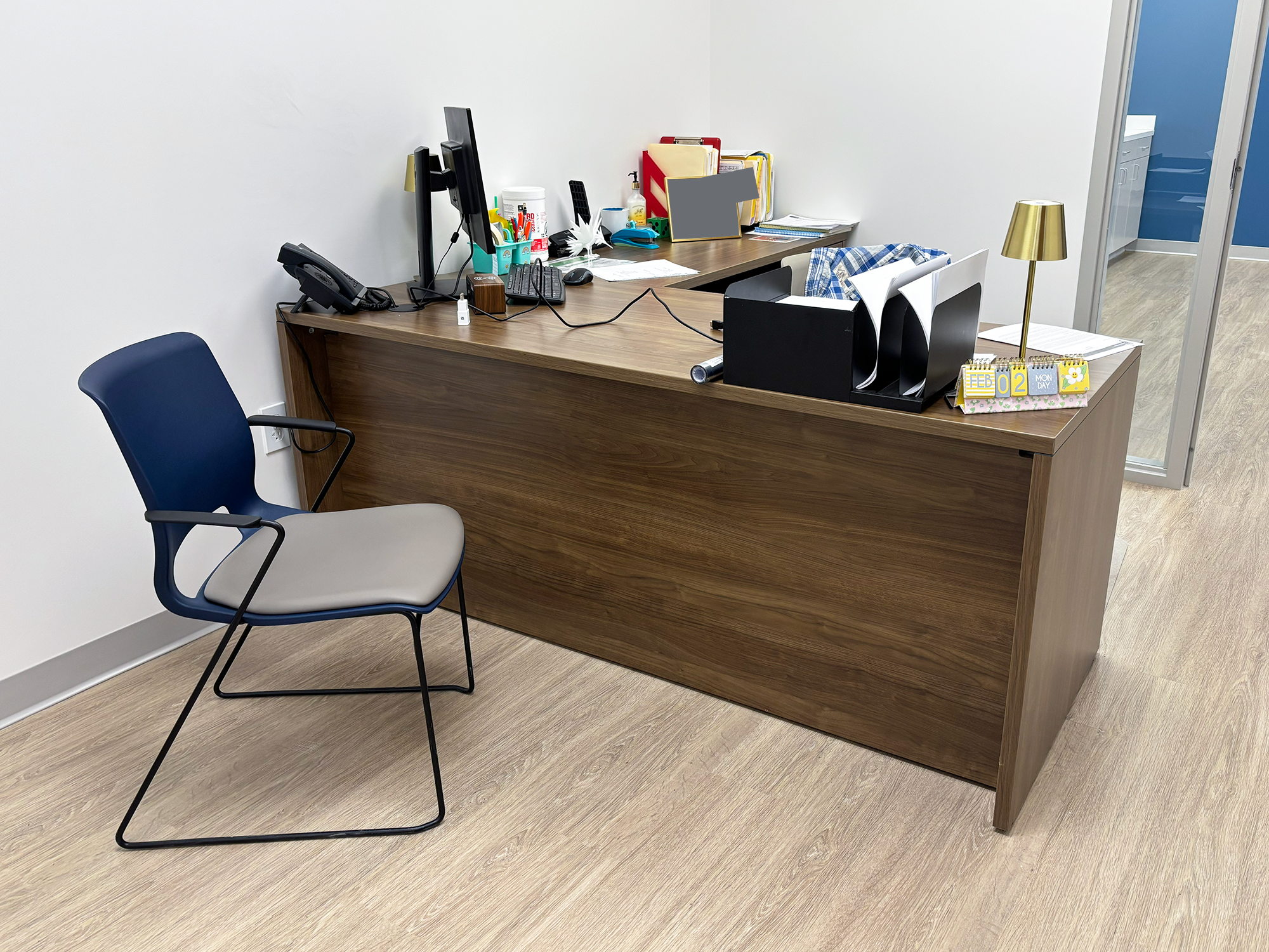 A desk and chair fill up a small office. The desk has a computer and other supplies on it